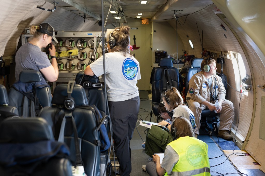 Looking down the length of a plane, several students and faculty stand by instruments, sit on the ground, and sit looking out the window.
