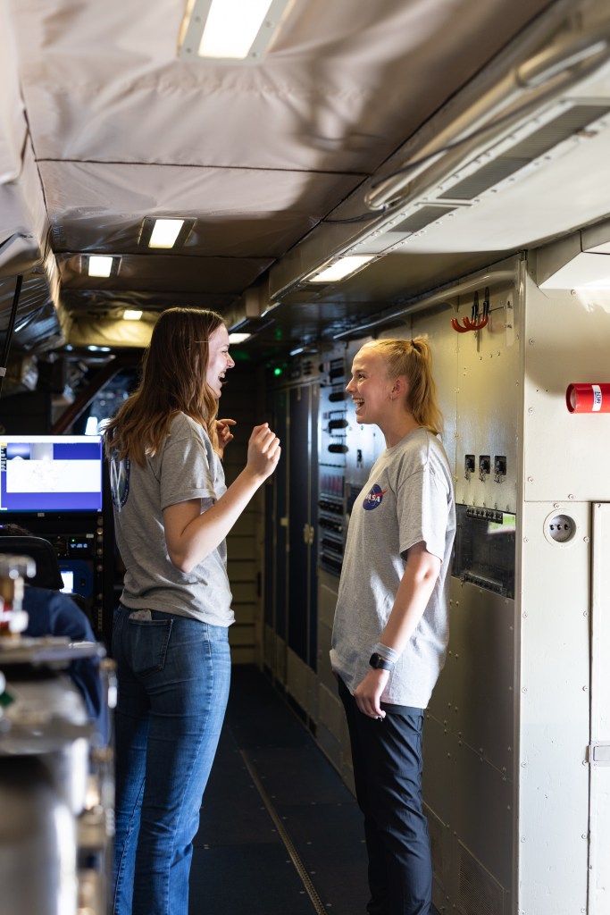 Two women in gray t-shirts stand talking and smiling in an aircraft hallway between instruments.