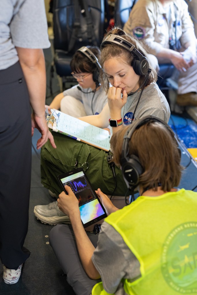 Three people sit on the ground wearing headphones, looking at data on a tablet and clipboard.