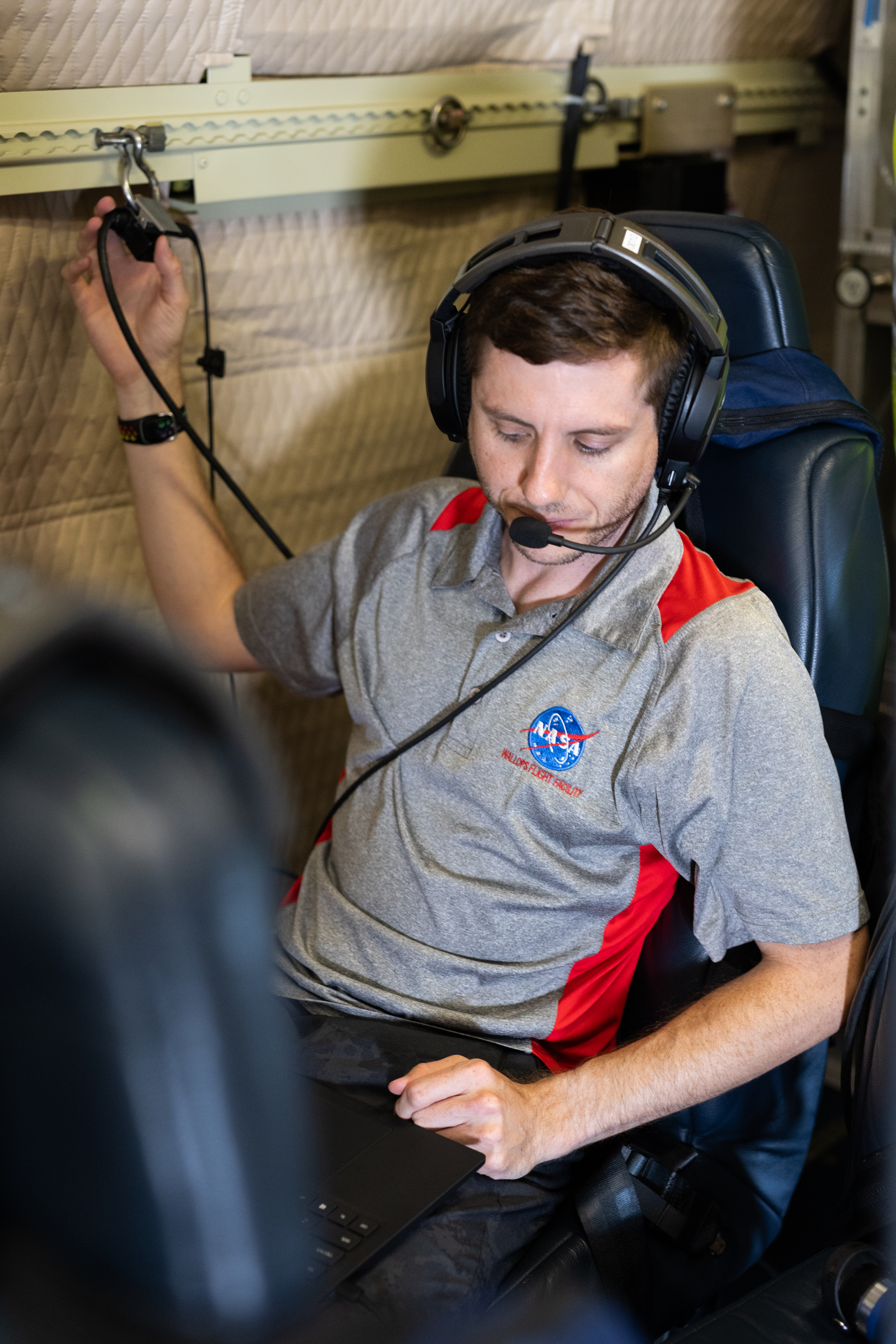 A man with short brown hair, wearing headphones and a gray shirt with a NASA logo sits on a blue seat working on a laptop.