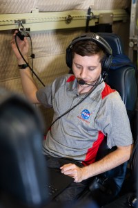 A man with short brown hair, wearing headphones and a gray shirt with a NASA logo sits on a blue seat working on a laptop.