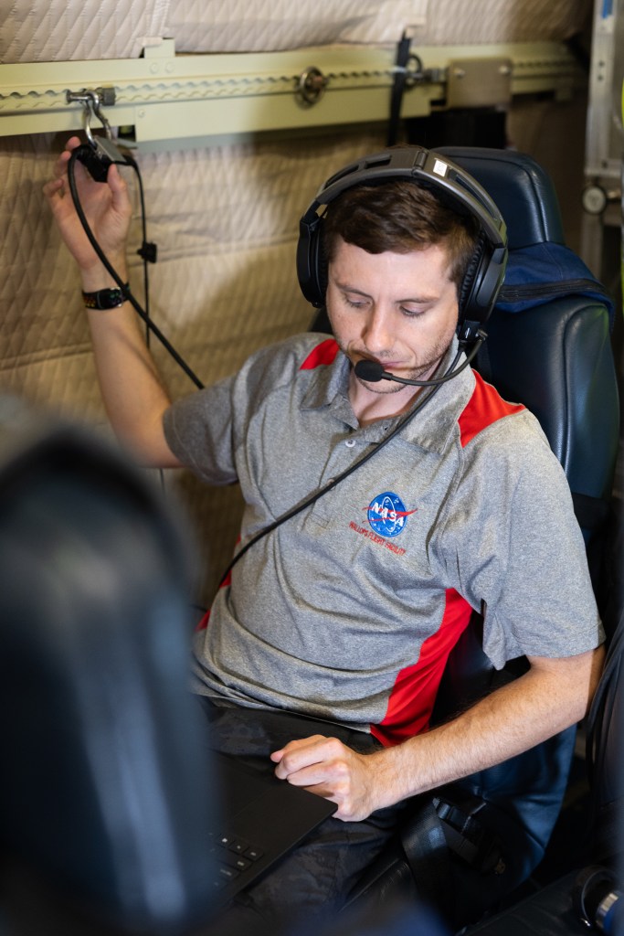 A man with short brown hair, wearing headphones and a gray shirt with a NASA logo sits on a blue seat working on a laptop.