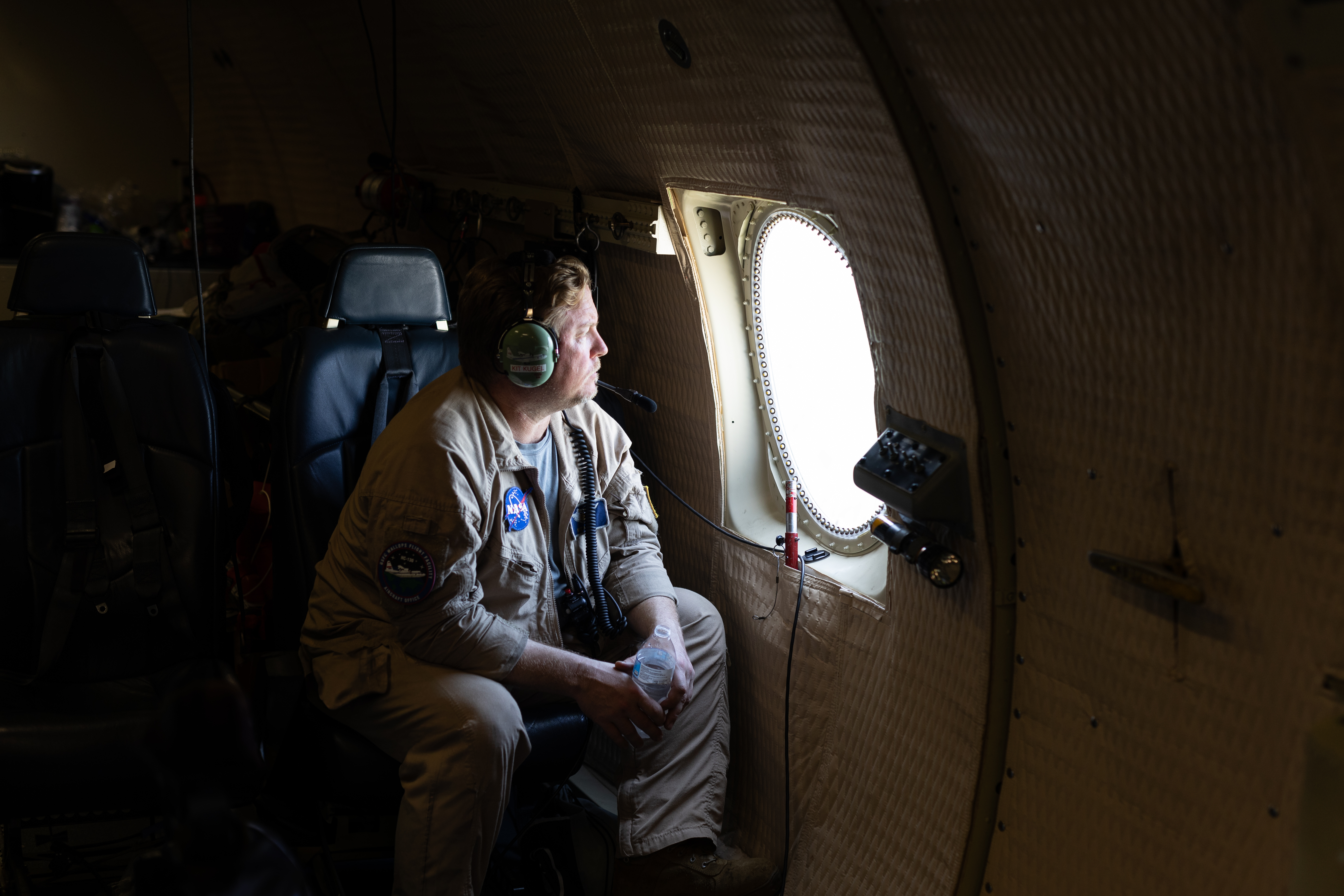 A man in a tan flight suit and headphones sits in an aircraft, peering out a small round window.