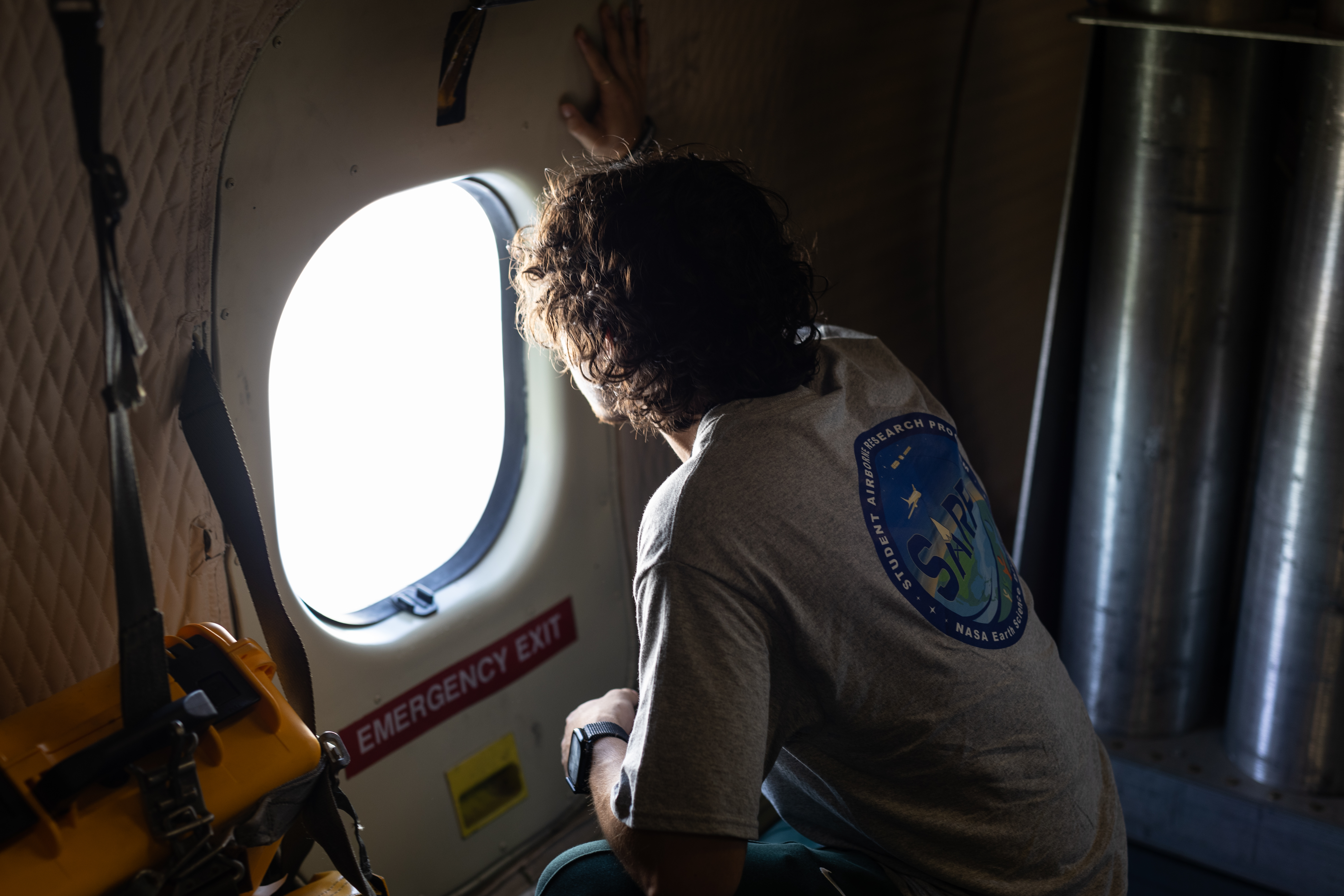 A student with curly hair, wearing a gray t-shirt with the SARP logo on the back, crouches on the floor of an aircraft peering out a small round window.