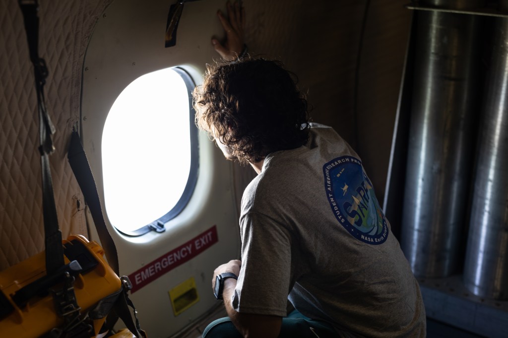 A student with curly hair, wearing a gray t-shirt with the SARP logo on the back, crouches on the floor of an aircraft peering out a small round window.