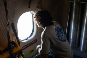 A student with curly hair, wearing a gray t-shirt with the SARP logo on the back, crouches on the floor of an aircraft peering out a small round window.