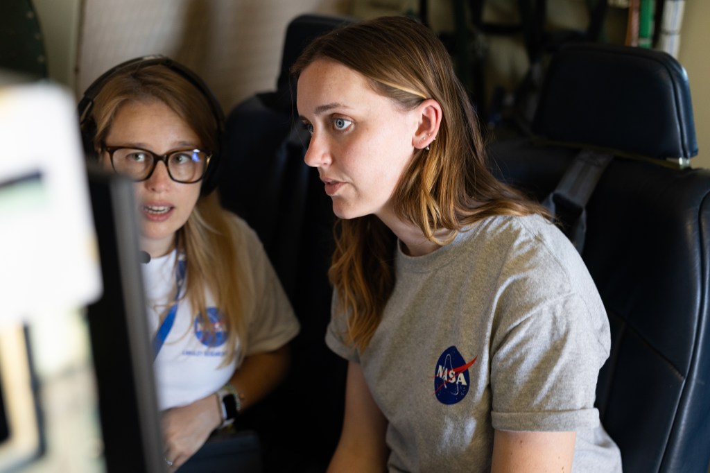 Two women in gray t-shirts with the NASA logo sit together looking at a screen.