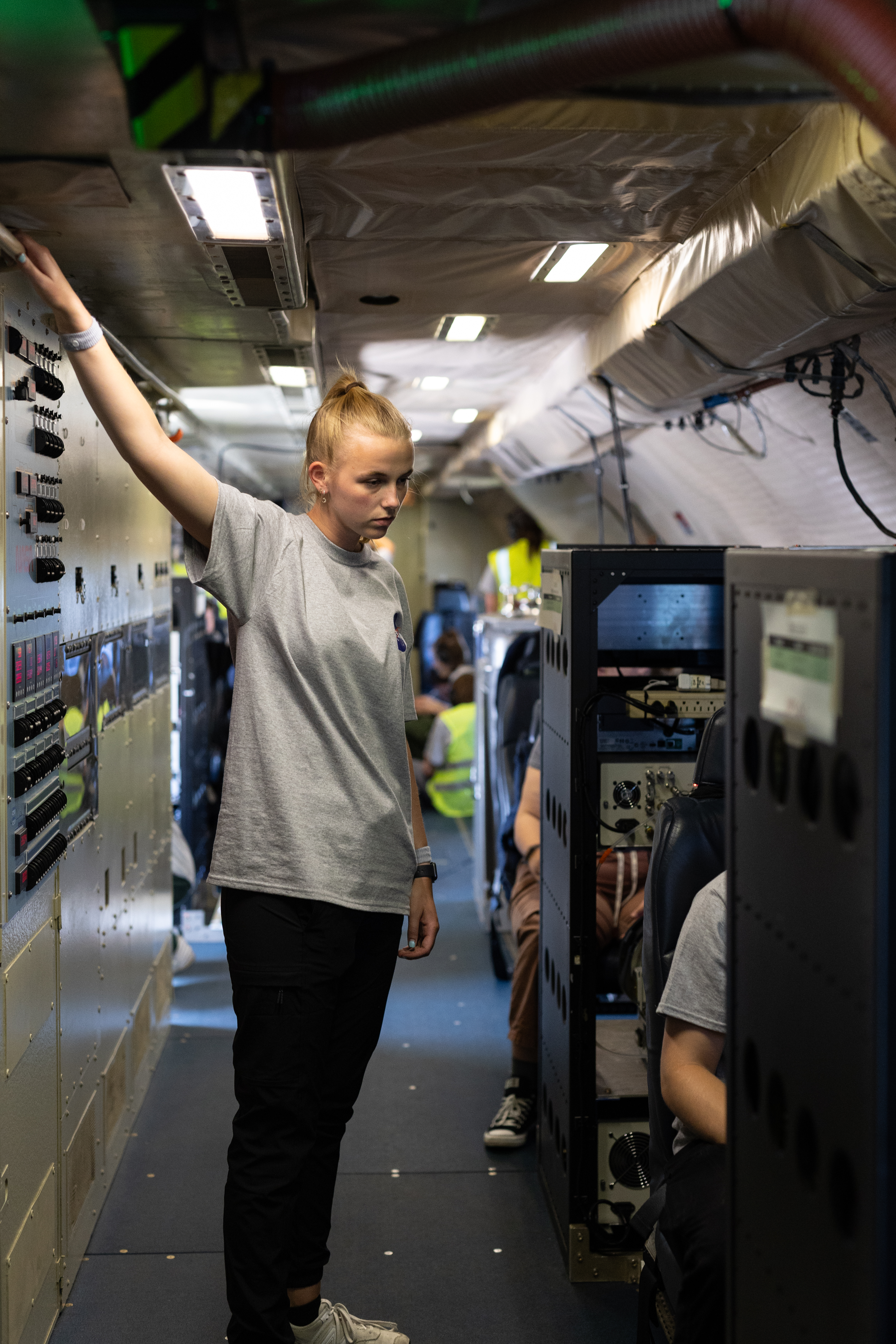 A student in a gray t-shirt and black pants stands on an aircraft looking at a dark gray box-shaped instrument.