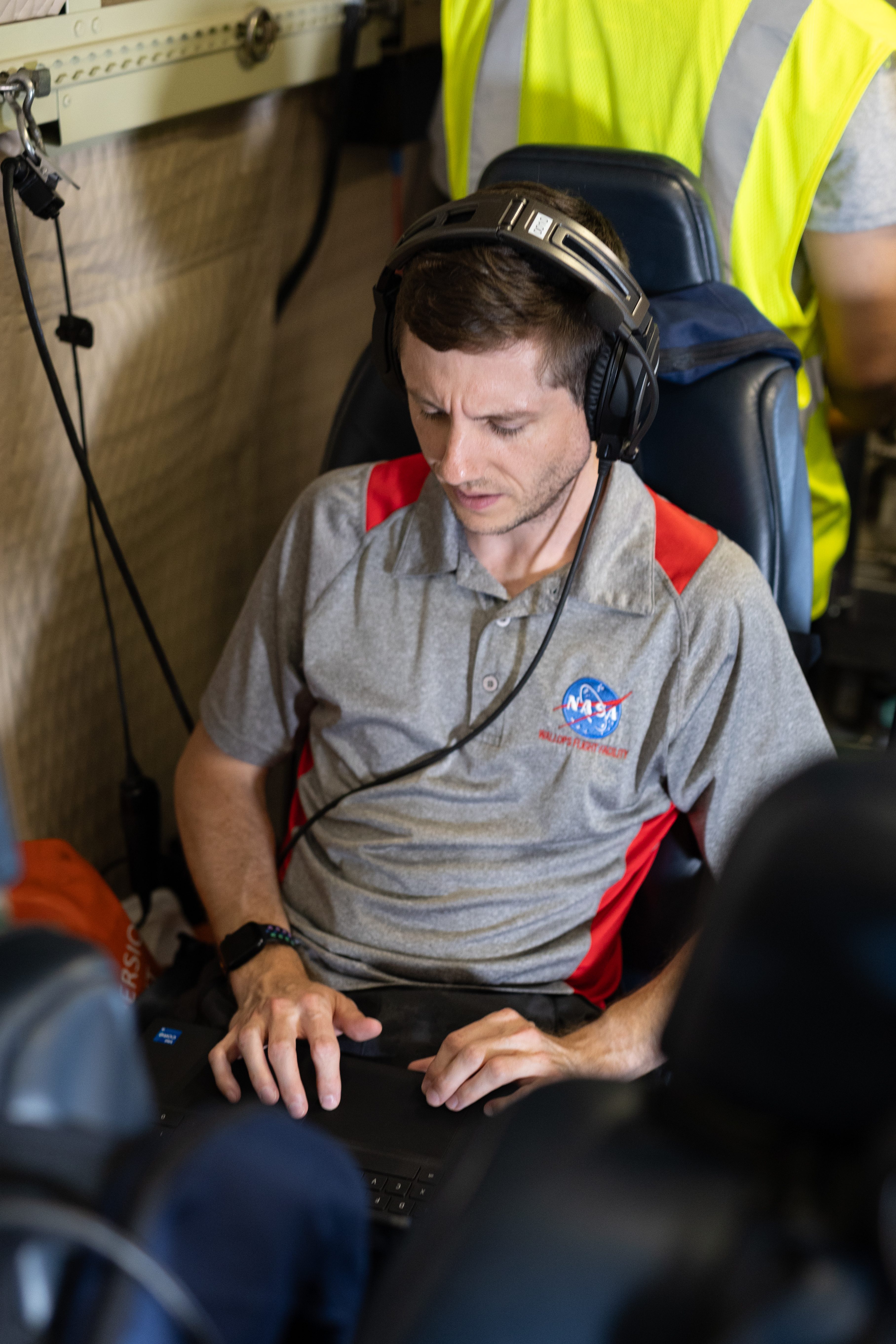 A man with short brown hair, wearing headphones and a gray shirt with a NASA logo sits on a blue seat working on a laptop.
