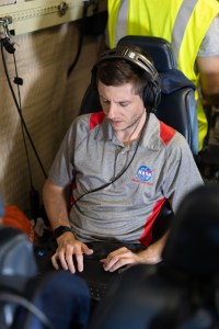 A man with short brown hair, wearing headphones and a gray shirt with a NASA logo sits on a blue seat working on a laptop.