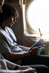A student with short, curly hair sits next to a small round window, looking down at a tablet.