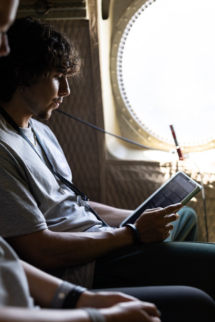 A student with short, curly hair sits next to a small round window, looking down at a tablet.