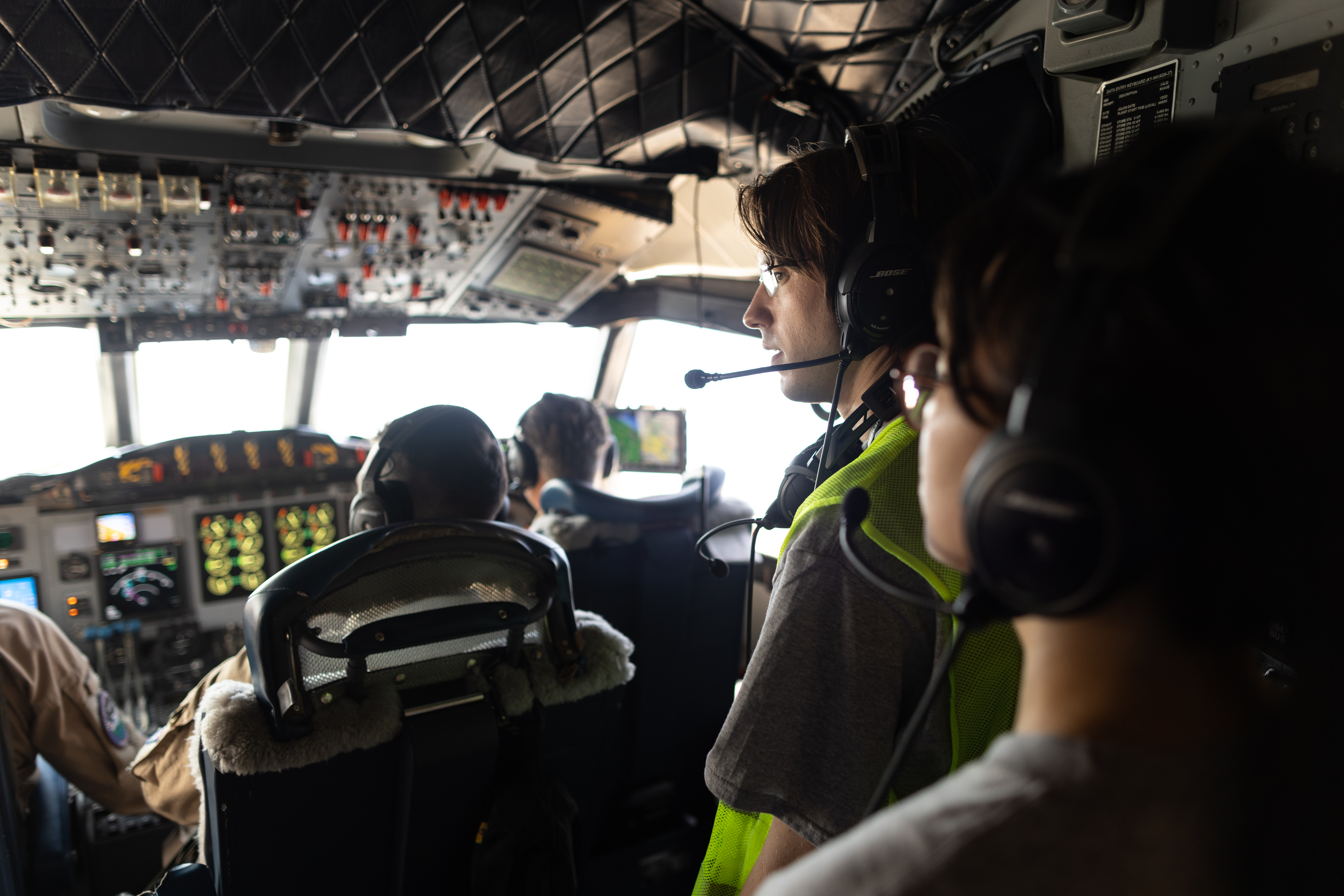 Two students with headphones stand in the back of a cockpit, watching three pilots in flight suits fly the plane.