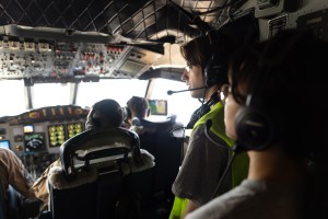 Two students with headphones stand in the back of a cockpit, watching three pilots in flight suits fly the plane.