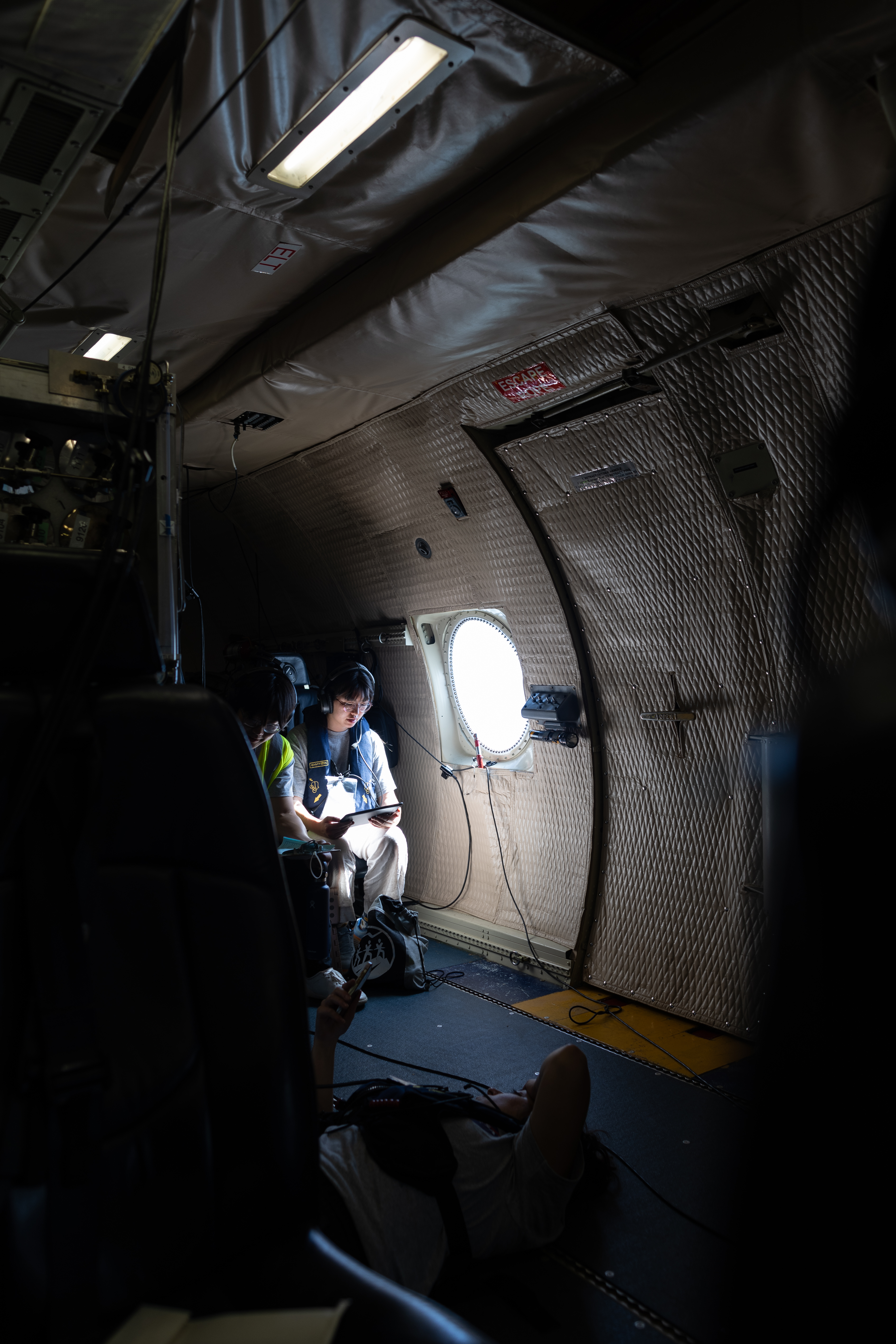 A student in a gray t-shirt and headphones sits in an aircraft next to a small round window, looking down at a tablet.