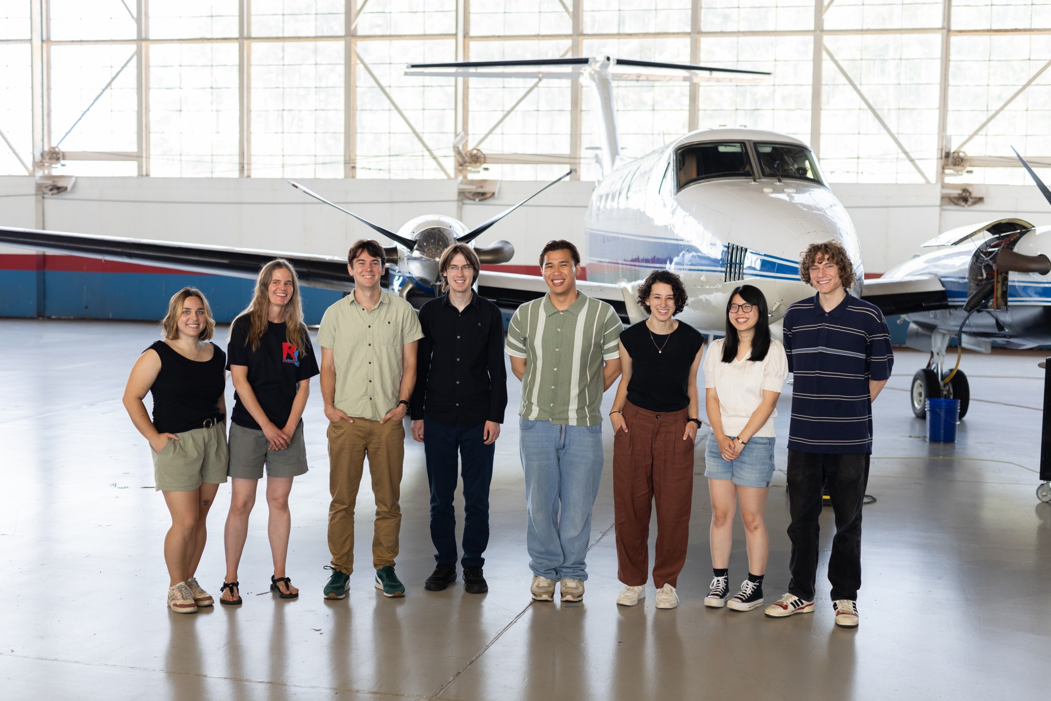 A group of eight people stand together inside a hangar with a somewhat shiny floor. In the background is a small white plane with a blue stripe, and large windows behind that.