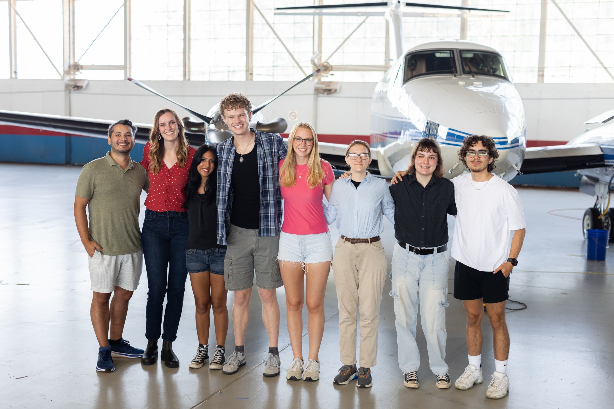 A group of eight people stand together inside a hangar with a somewhat shiny floor. In the background is a small white plane with a blue stripe, and large windows behind that.
