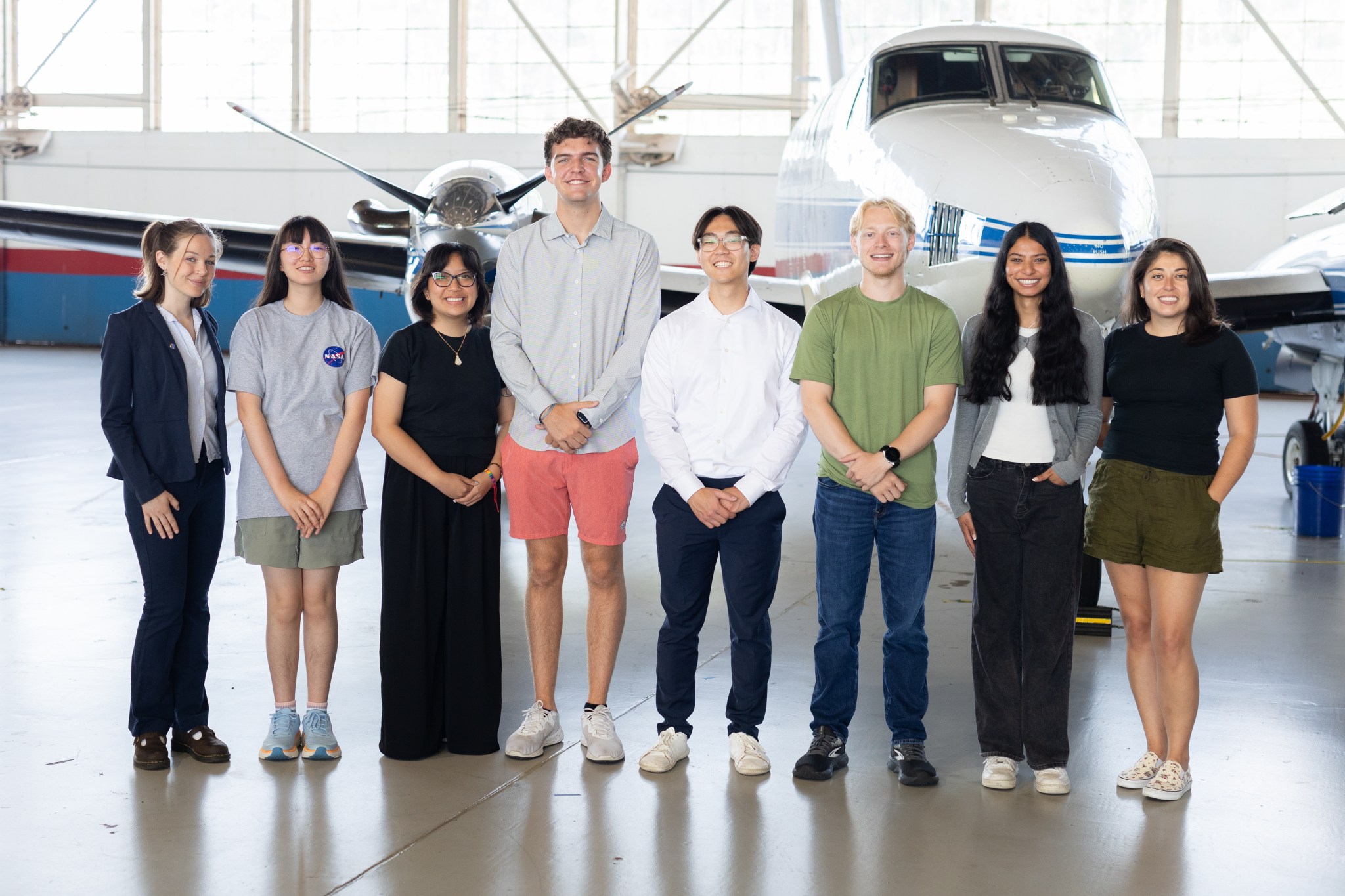A group of eight people stand together inside a hangar with a somewhat shiny floor. In the background is a small white plane with a blue stripe, and large windows behind that.