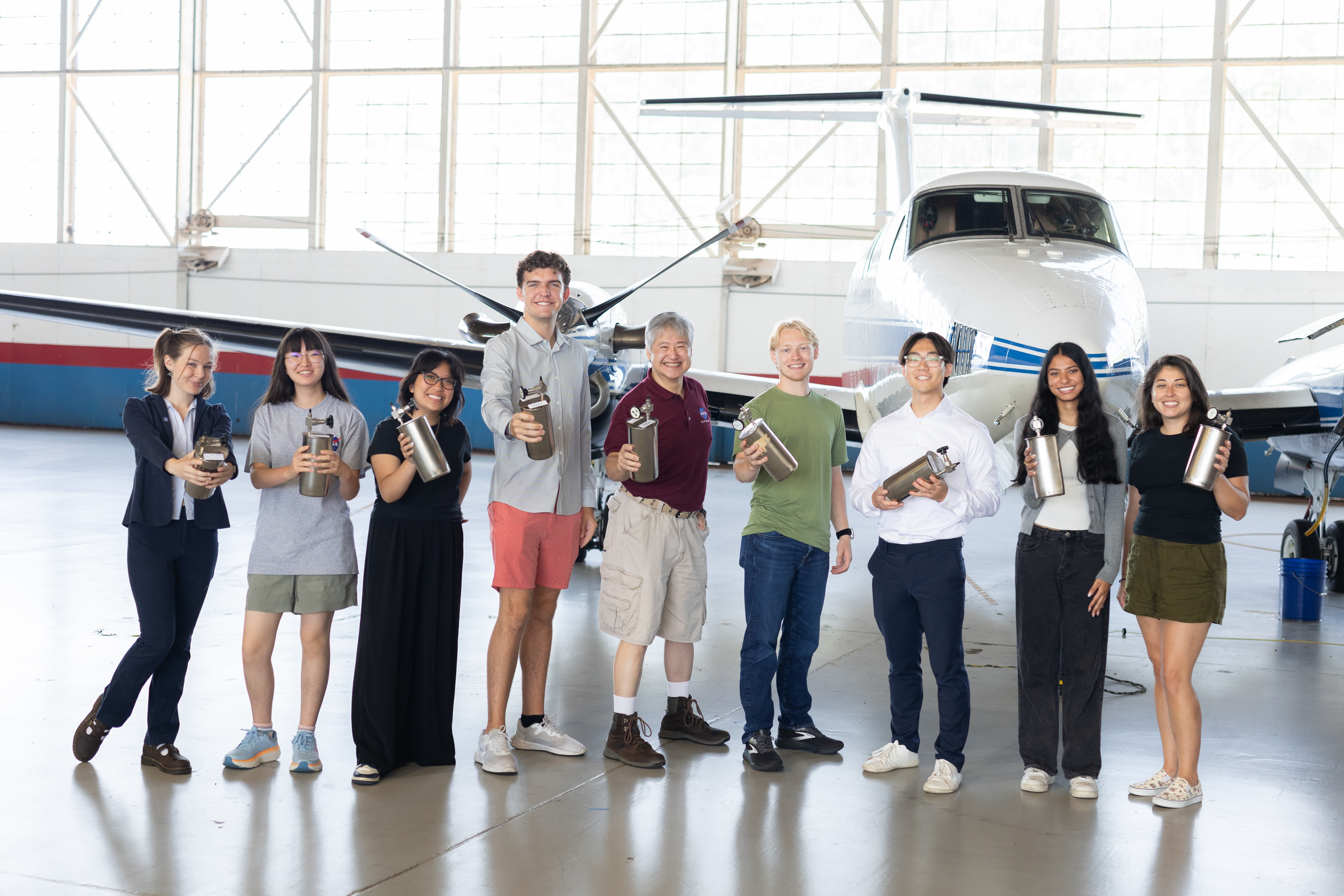 A group of eight people stand together inside a hangar with a somewhat shiny floor, holding small silver canisters a bit larger than a thermos. In the background is a small white plane with a blue stripe, and large windows behind that.
