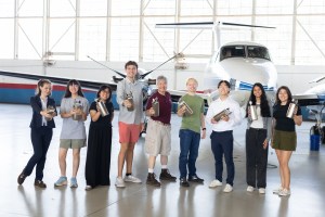A group of eight people stand together inside a hangar with a somewhat shiny floor, holding small silver canisters a bit larger than a thermos. In the background is a small white plane with a blue stripe, and large windows behind that.