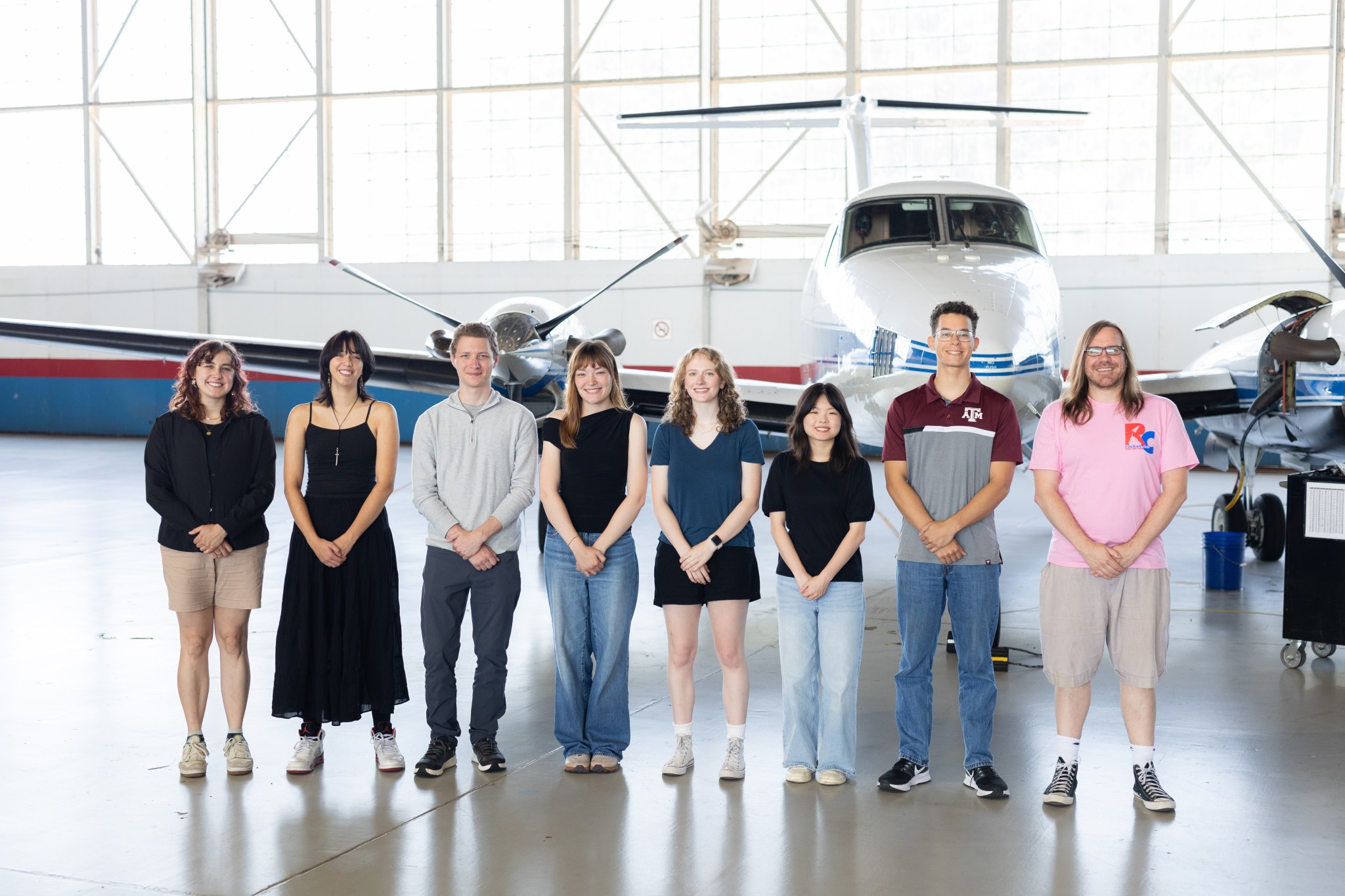 A group of eight people stand together inside a hangar with a somewhat shiny floor. In the background is a small white plane with a blue stripe, and large windows behind that.