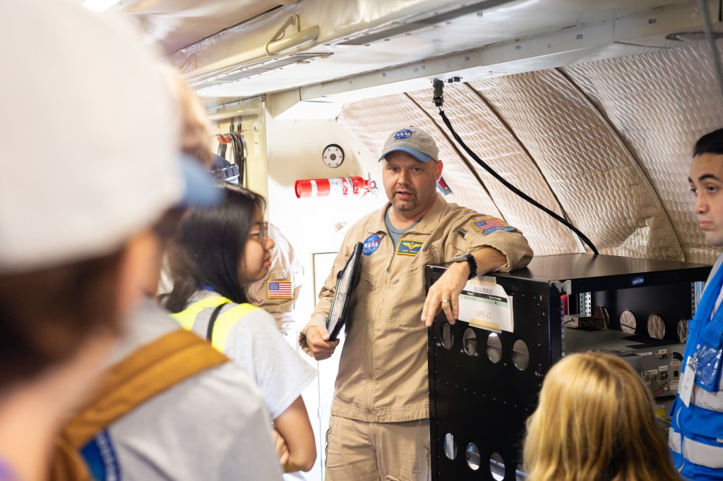 A man in a baseball cap and tan flightsuit holding a clipboard speaks to a group of students with their backs to the camera.