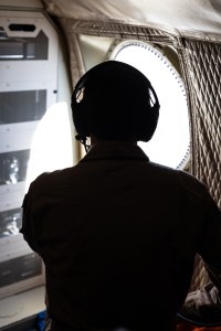 A silhouette of a man wearing headphones staring out a round plane window.