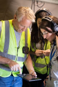 A man with white hair wearing a high-viz vest shows two female students readouts on a small tablet.