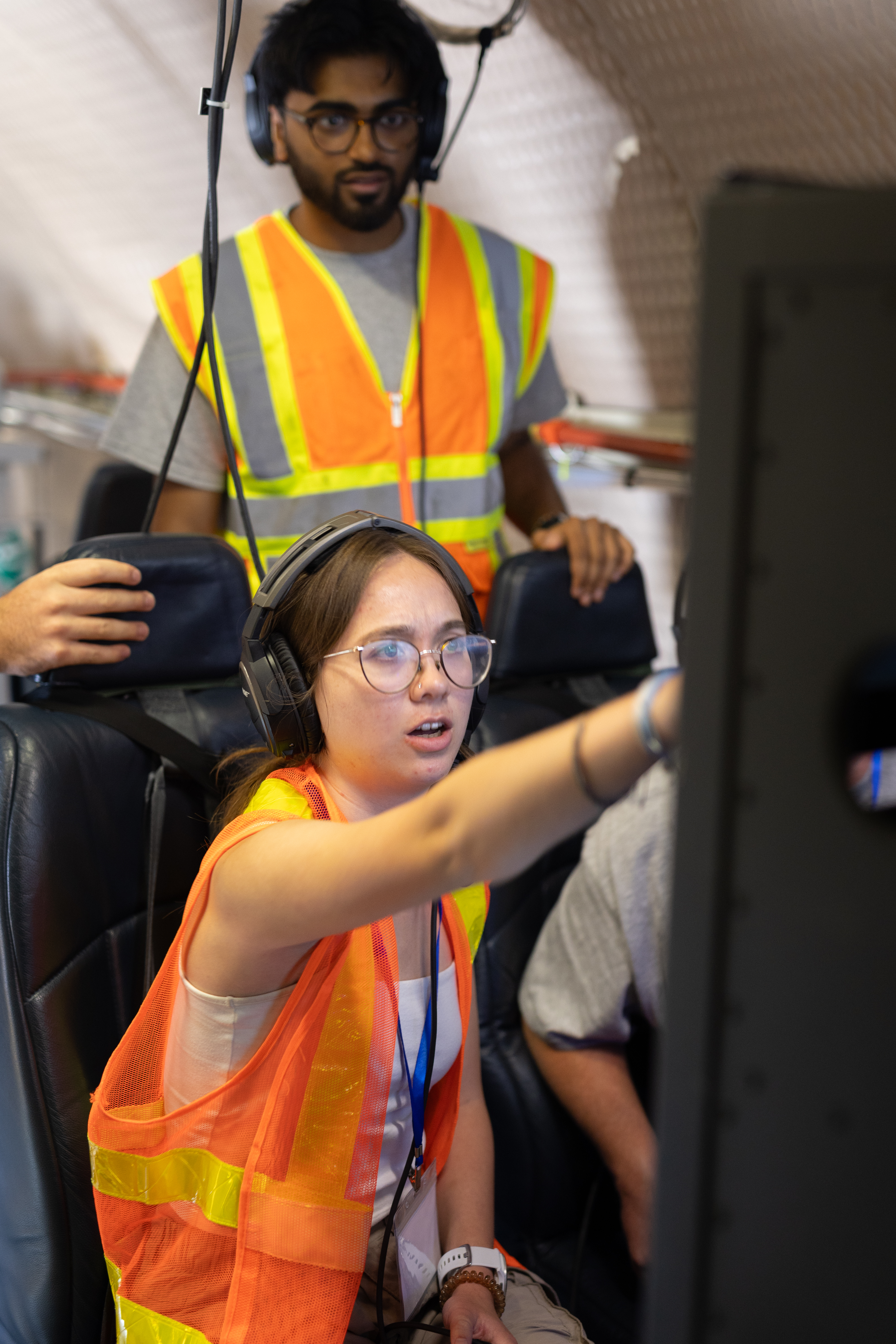Two students in orange high-viz vests and headphones look at a computer screen. The male student in the back is standing, and the female student in front is sitting in the aircraft seat and pointing at the screen.