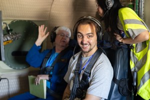 A student in a gray t-shirt and headphones smiles at the camera. In the background, an instructor in all blue with white hair explains something to another student in a yellow high-viz vest.