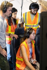 Four students wearing headphones, three with orange high-viz vests on, gather around a screen.