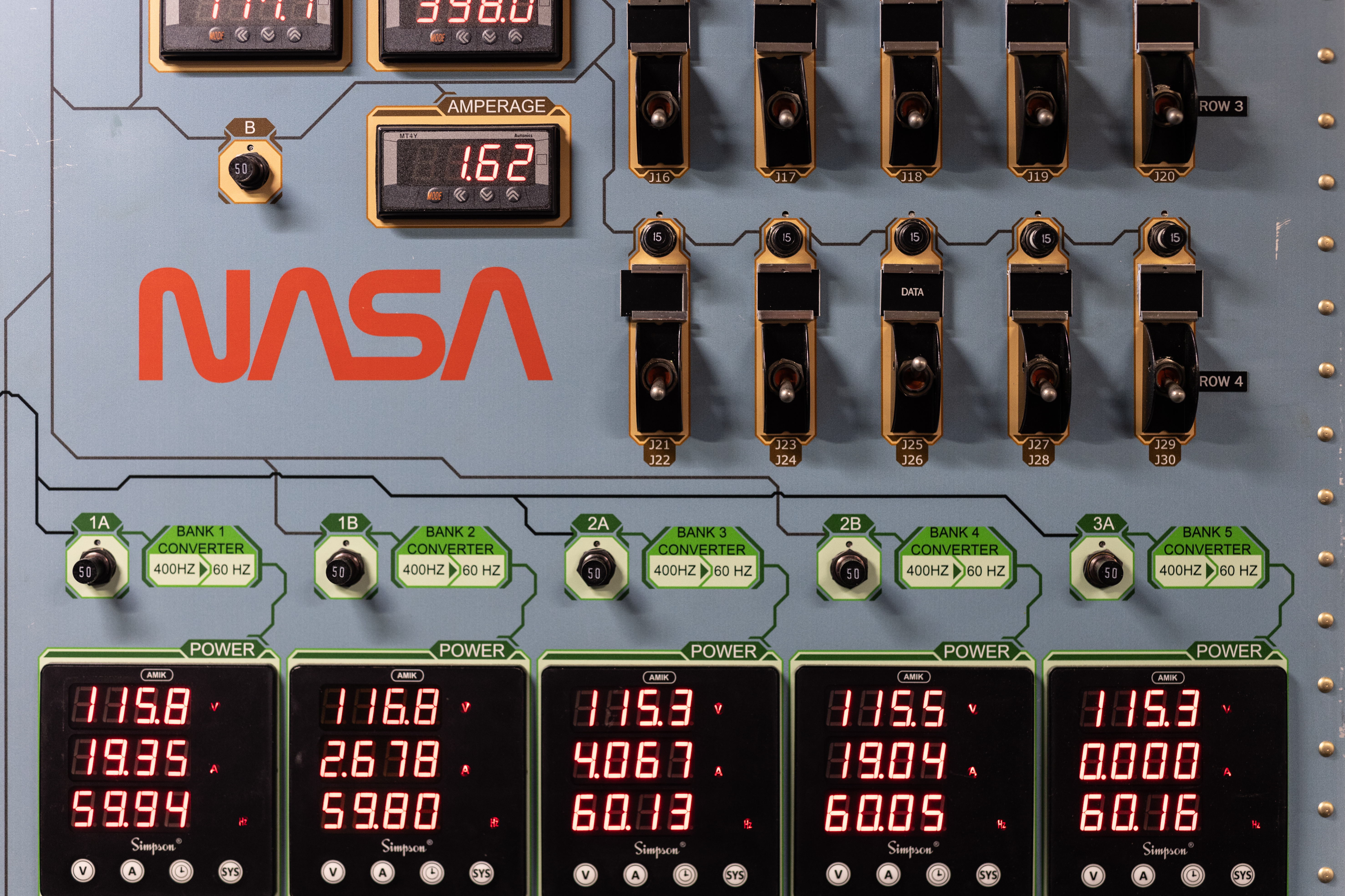 A gray pannel covered in switches and digital displays with red numbers, with the NASA worm logo in red on the left.