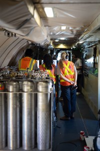 Looking down the body of a plane, three students wearing headphones and orange high-viz vests stand with their back to the camera, looking at two screens. In the foreground is a group of tall silver cylinders.