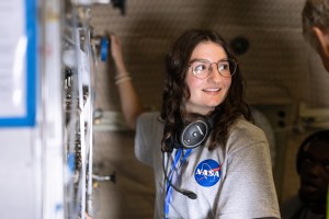 A female student with dark brown hair and glasses, wearing a gray shirt with the NASA logo on it, adjusts the valves on a large gray machine.