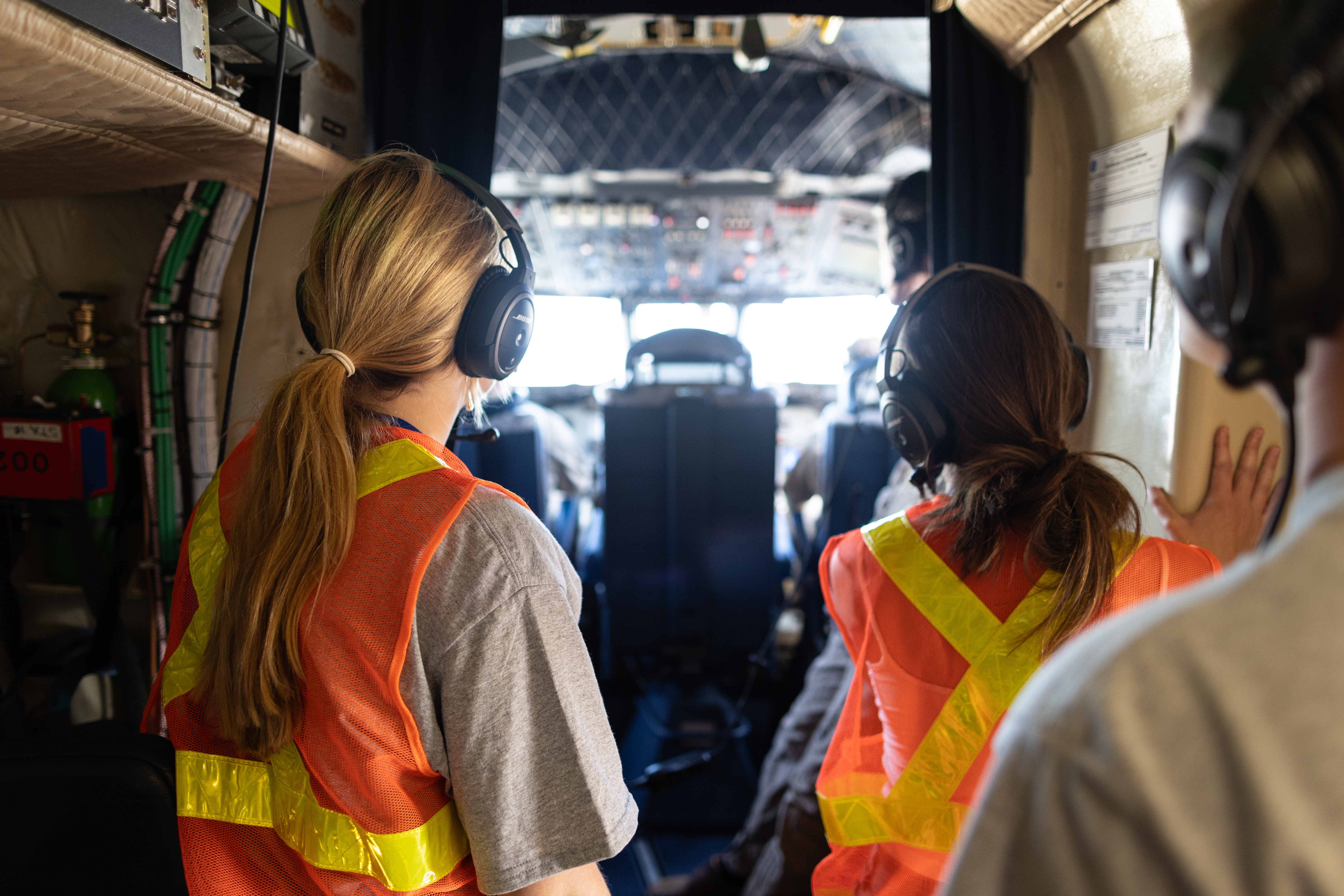 Two female students wearing headphones and orange high-viz vests peer into a cockpit.