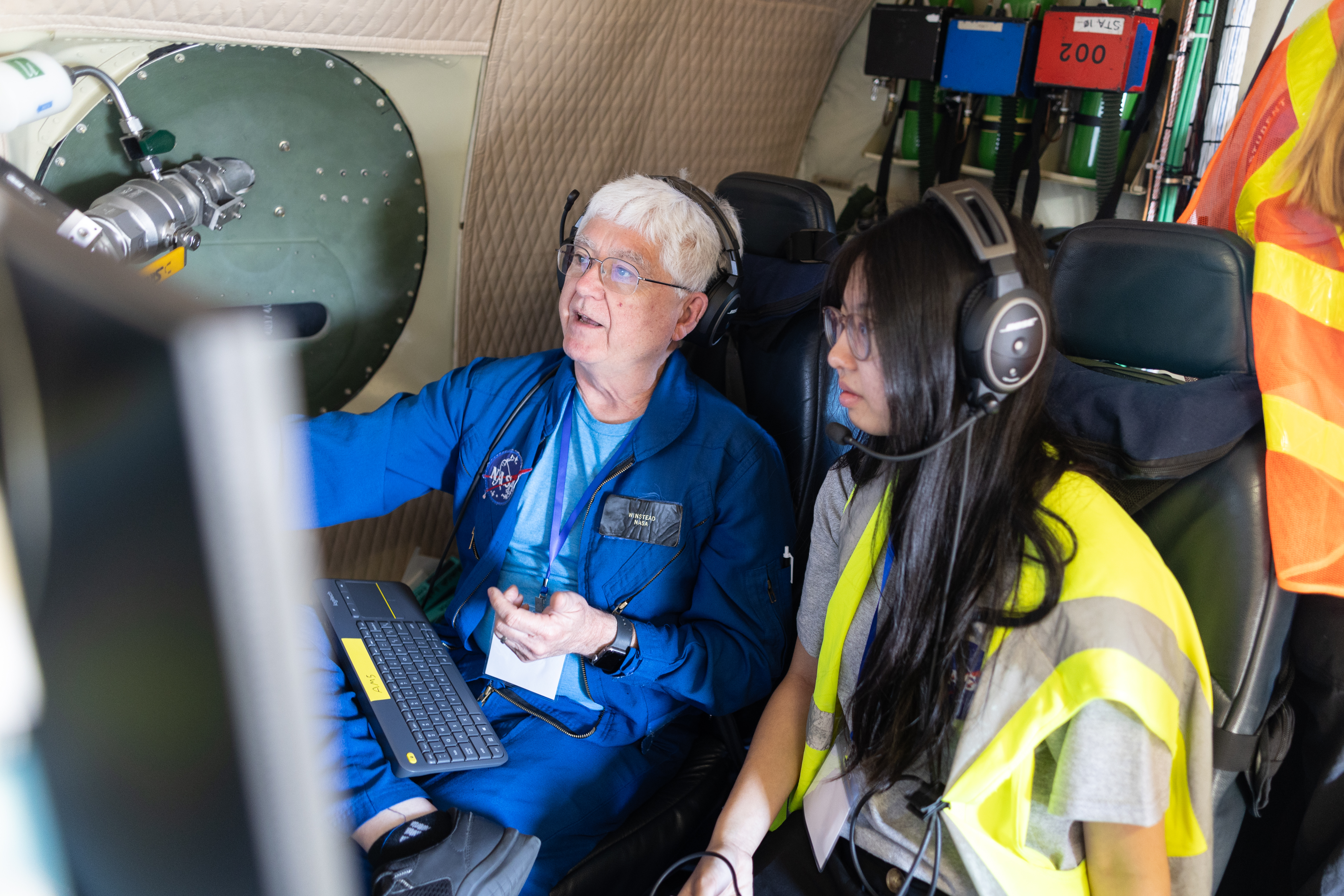 An instructor with white hair and a blue flightsuit points at a screen, explaining data to a student sitting next to him. The student has long black hair, headphones, and a yellow high-viz vest.