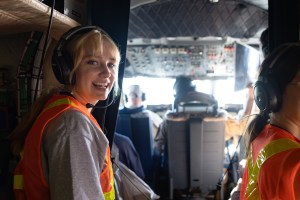 A female students with blonde hair and bangs and wearing headphones and an orange high-viz vest smiles at the camera, with a plane cockpit in the background.