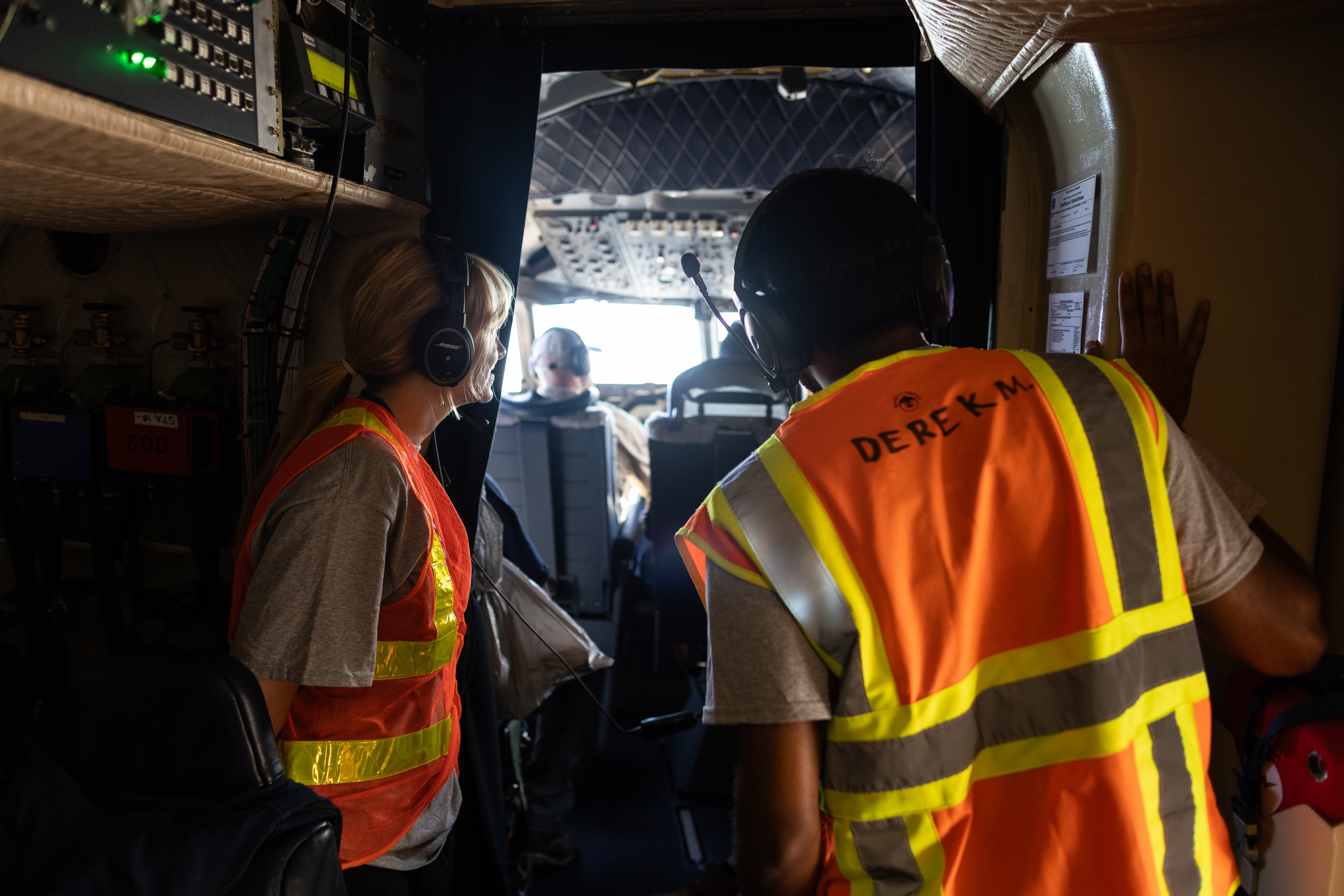 Two students wearing headphones and orange high-viz vests peer into a cockpit.