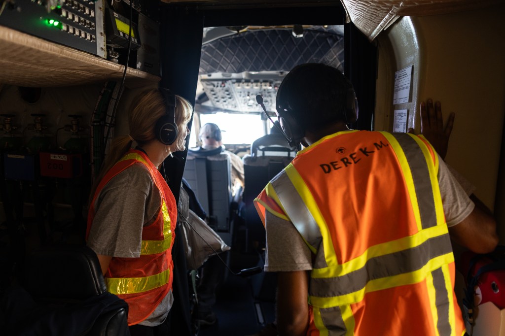 Two students wearing headphones and orange high-viz vests peer into a cockpit.