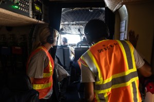 Two students wearing headphones and orange high-viz vests peer into a cockpit.