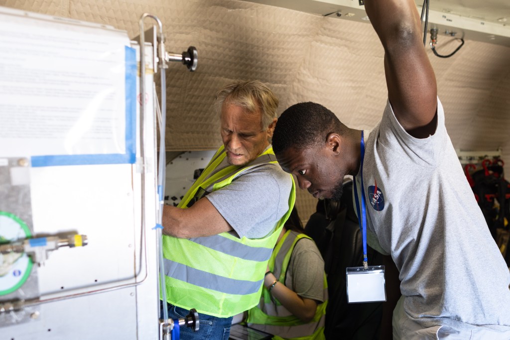 A man with white hair and a yellow high-viz vest stands next to a student in a gray t-shirt, as they both peer intently at a gray instrument in front of them.