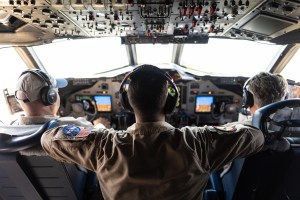 Three men in headphones sit in a cockpit with their backs to the camera, watching the displays.
