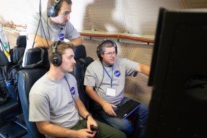 Two men in gray shirts and headphones sit looking at a screen. A student dressed the same stand behind them, leaning on the back of the chairs.
