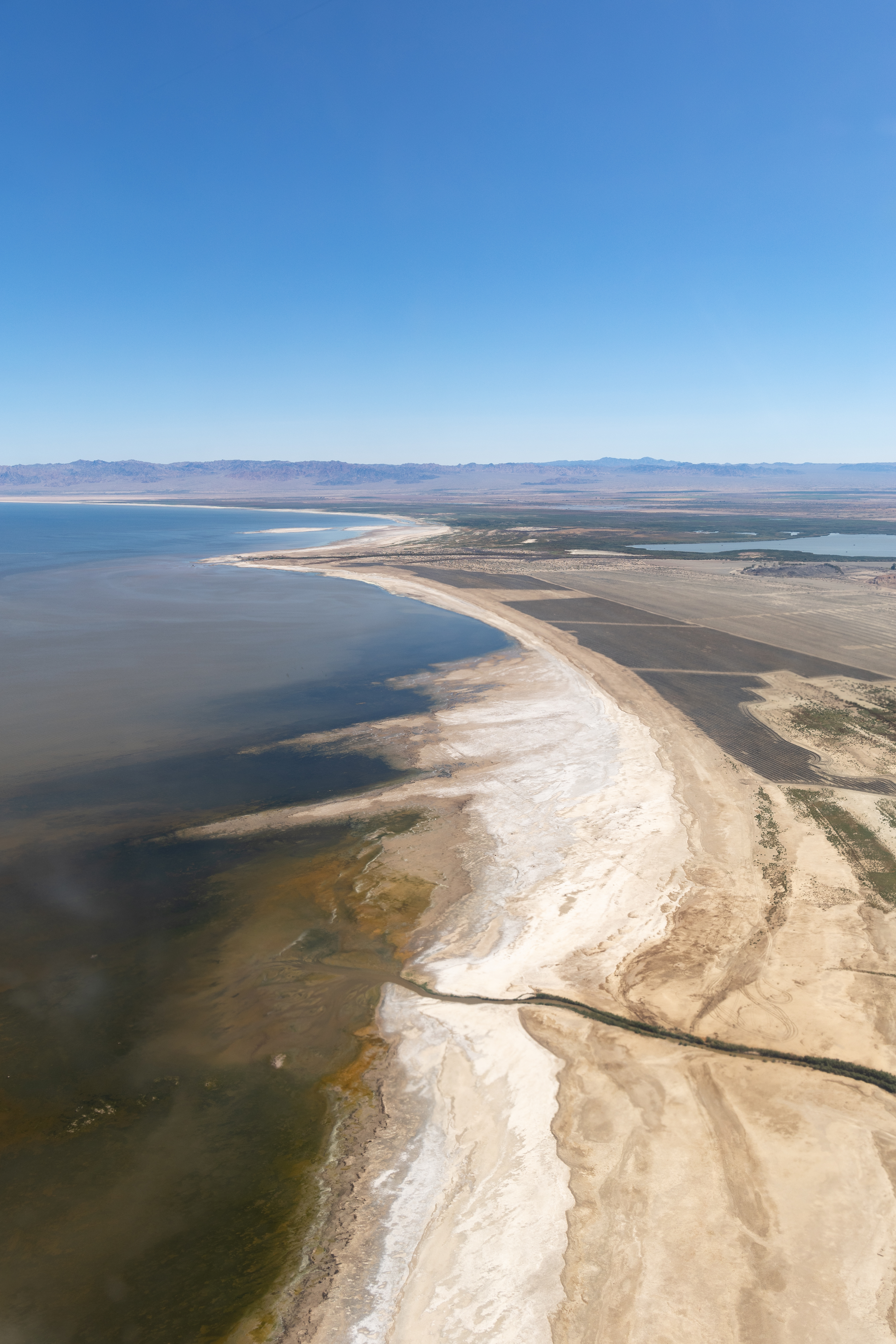 A high view of a lakeshore shows variegated blueish brown water, on a coast with white and tan sand.