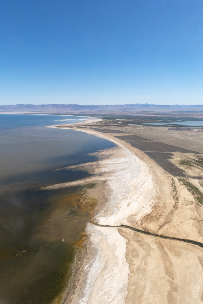 A high view of a lakeshore shows variegated blueish brown water, on a coast with white and tan sand.
