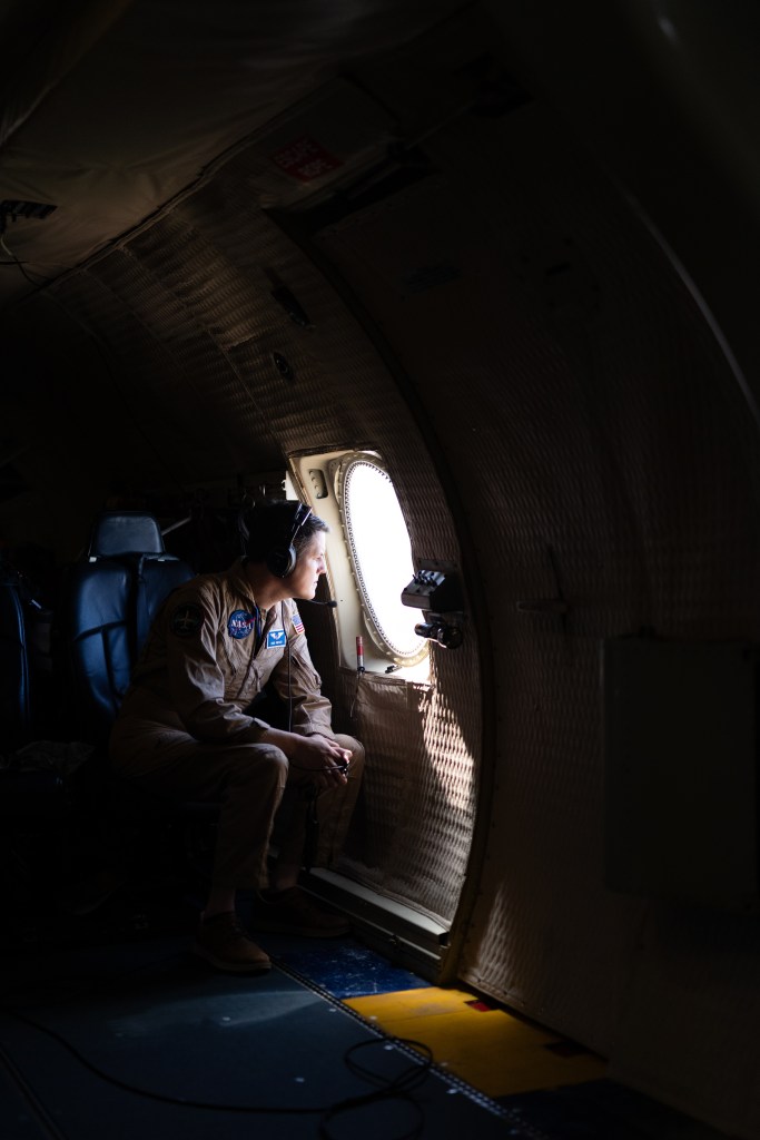 A man in a tan flight suit and headphones sits in an aircraft, peering out a small round window.