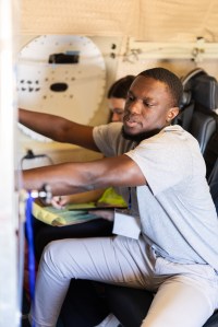 A student with short black hair and a gray t-shirt sits in an aircraft chair, adjusting two valves on the machine in front.