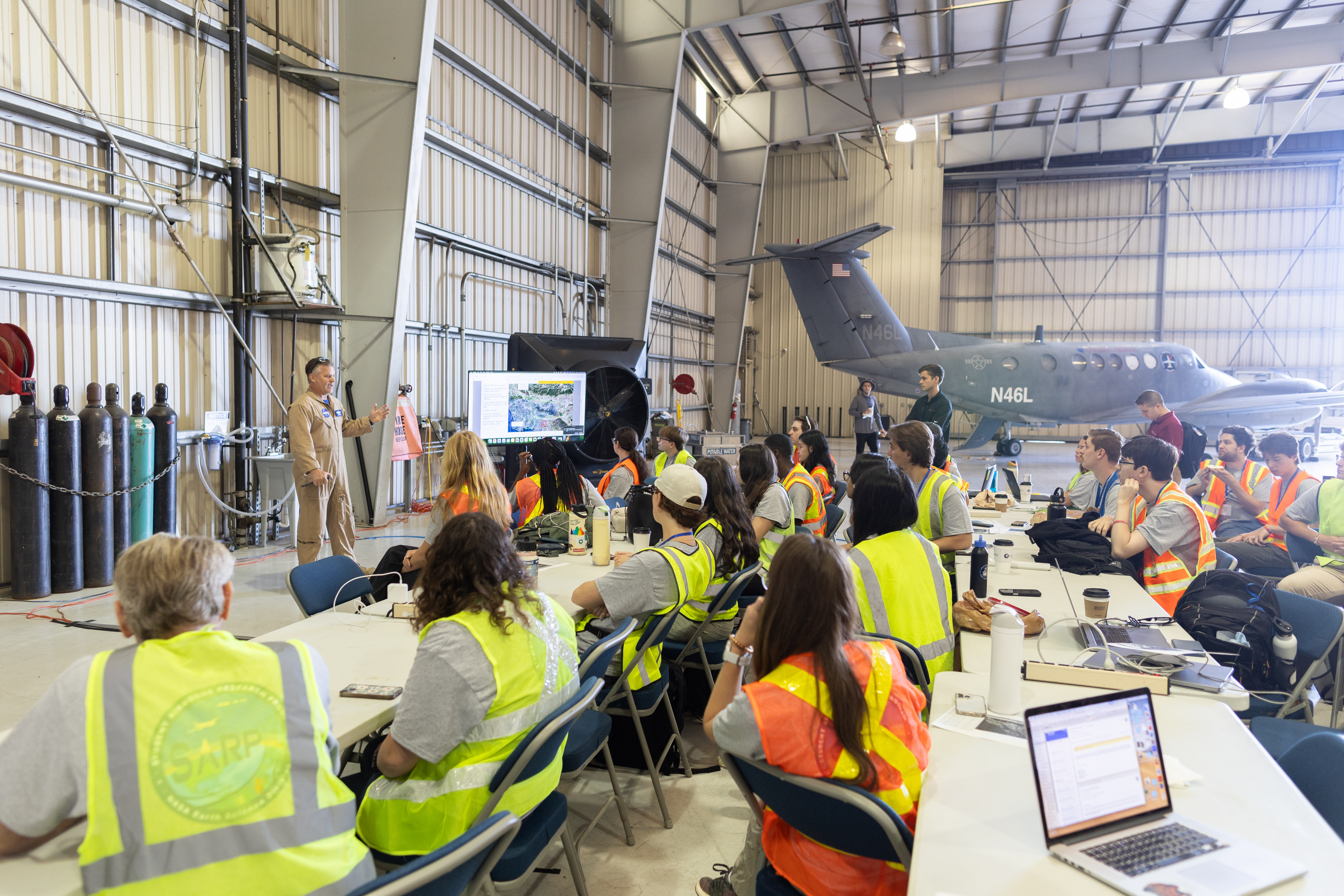 Students in high-viz vests sit around white tables with their backs to the camera, listenting to a man in a tan flight suit standing next to a portable screen with a map graphic on it.