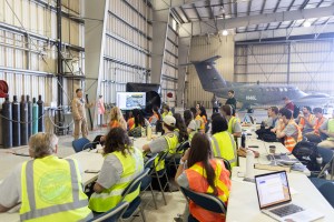 Students in high-viz vests sit around white tables with their backs to the camera, listenting to a man in a tan flight suit standing next to a portable screen with a map graphic on it.