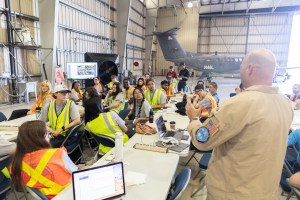Students in high-viz vests sit around white tables, listenting to a bald man in a tan flight suit standing in the bottom left corner of the frame.